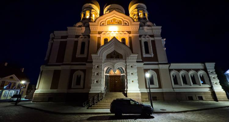 Façade d'église orthodoxe illuminée avec dômes capturée la nuit, voiture garée solitaire devant.