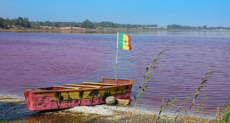 Un bateau en bois coloré arborant le drapeau sénégalais repose sur les eaux rose vif du lac Retba sous un ciel dégagé.