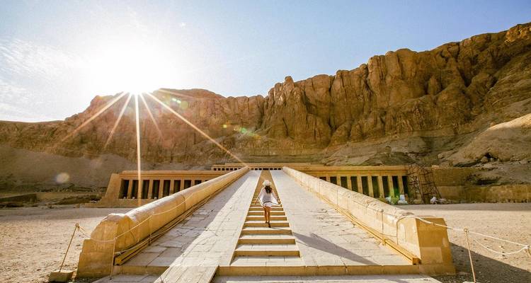 Traveller ascending the ramp toward the Temple of Hatshepsut with a dramatic sunburst over desert cliffs.