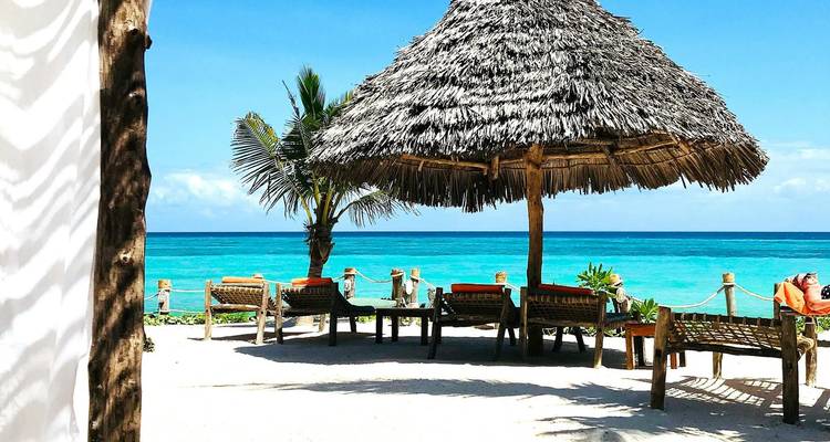 Idyllic white-sand beach with turquoise water, palm tree and thatched umbrella over lounge chairs.