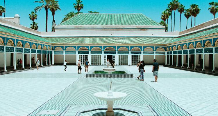 Elegant tiled courtyard with central fountain and green-roofed palace buildings, visitors walking around.