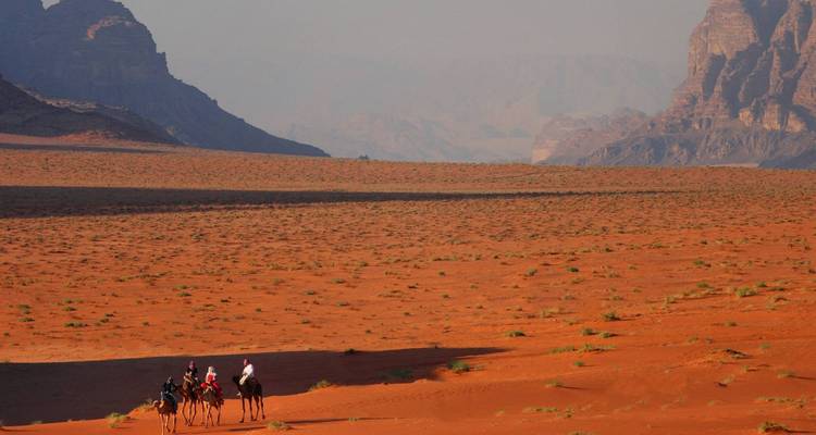 Tres viajeros en camellos caminan a través de la inmensa explanada de arena roja de Wadi Rum con formaciones rocosas imponentes a la distancia.