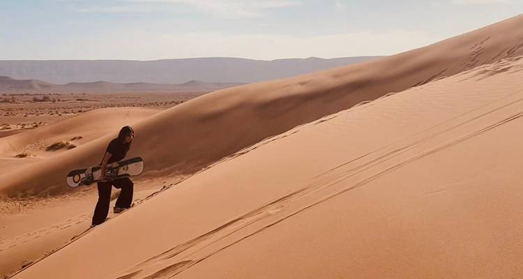 Traveller climbs steep sand dune carrying a sandboard in golden afternoon light.