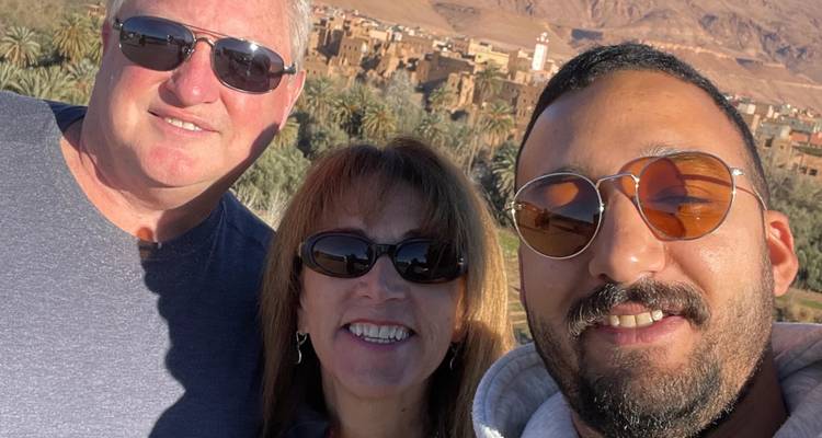 Selfie of three travellers with an oasis of palm trees and mud-brick village in the background.