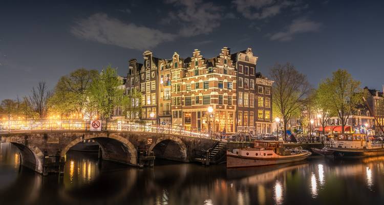 Un classique pont de canal d'Amsterdam et des maisons à pignon scintillent avec des illuminations nocturnes reflétées dans l'eau.