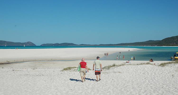 Plage de sable blanc étendue avec des eaux peu profondes turquoise où les visiteurs se promènent et prennent des bains de soleil sous un ciel bleu dégagé.