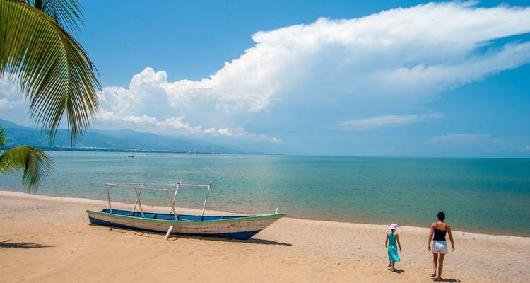 Woman and child walk along sandy Lake Tanganyika beach next to a wooden boat and palm frond.