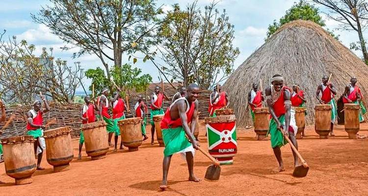 Burundi drummers in red and green attire beat large wooden drums beside a thatched hut.