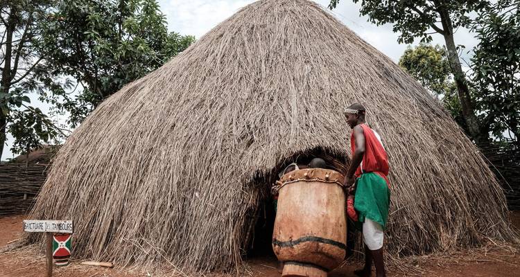 Man in traditional dress stands beside a huge drum at the entrance of a conical thatched hut.