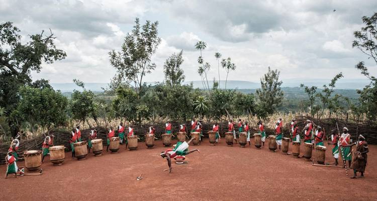Semi-circular formation of Burundian drummers performing outdoors on red soil with green hills behind.