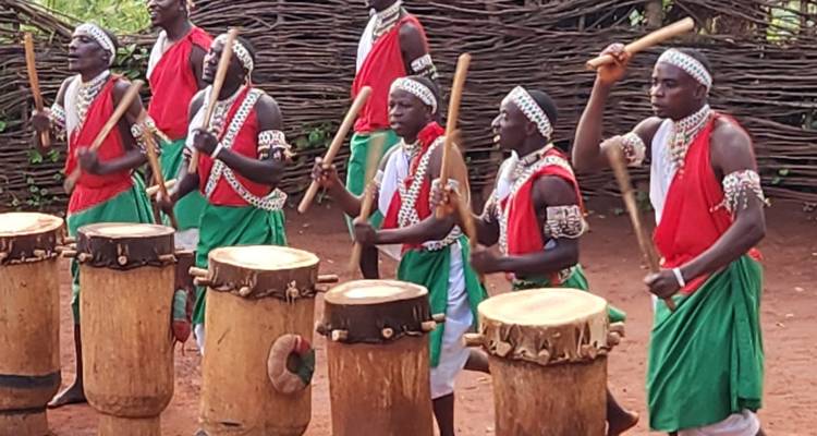 Line of Burundian drummers in red and green attire rhythmically beating tall wooden drums.