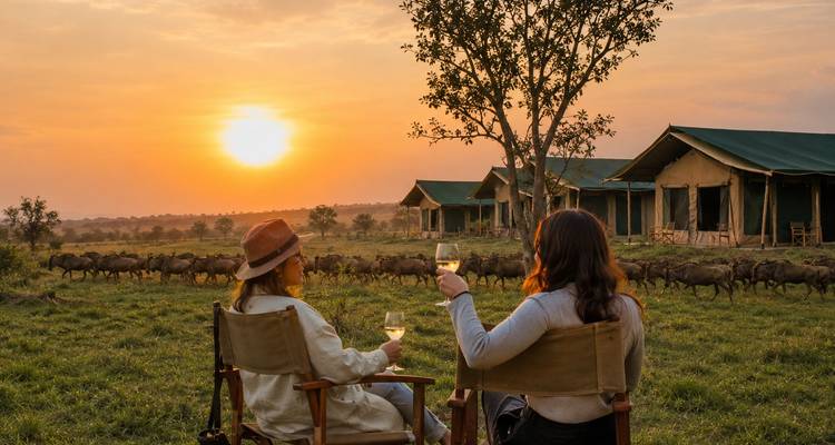 Two women toast wine at sunset while wildebeest graze near canvas safari tents in the distance.