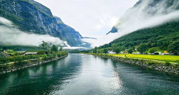 Rustige rivier die door een steil, mistaangevuld fjordvallei stroomt, omzoomd met groen.