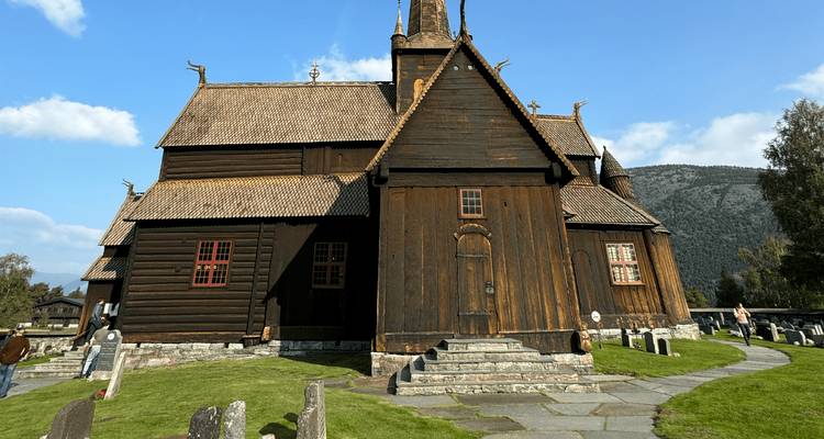 Historische houten staafkerk met een met gras bedekt dak van het schip tegen blauwe lucht en heuvels.