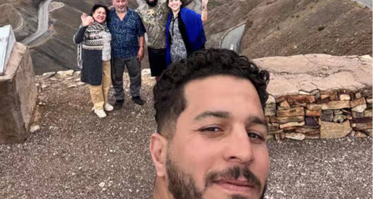 Un homme prend un selfie avec des amis qui font des gestes de la main sur un col de montagne surplombant des routes sinueuses.
