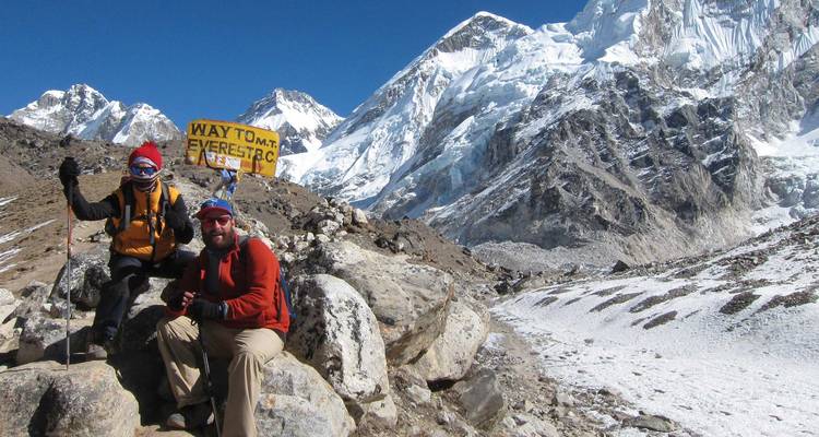 Dos trekkers descansan junto a un letrero amarillo que dice 'Way to Mt. Everest BC' en medio de picos nevados.