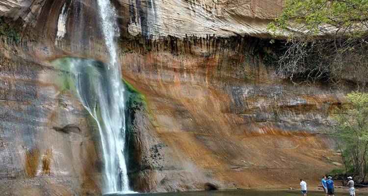 Eine kleine Gruppe steht am Fuße eines hohen Wüstenwasserfalls, der über mehrfarbiges Gestein hinabstürzt.