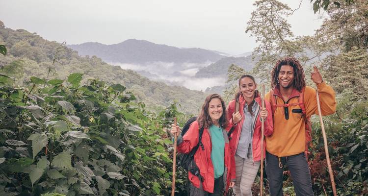 Three trekkers smile and gesture with misty forested hills behind them.