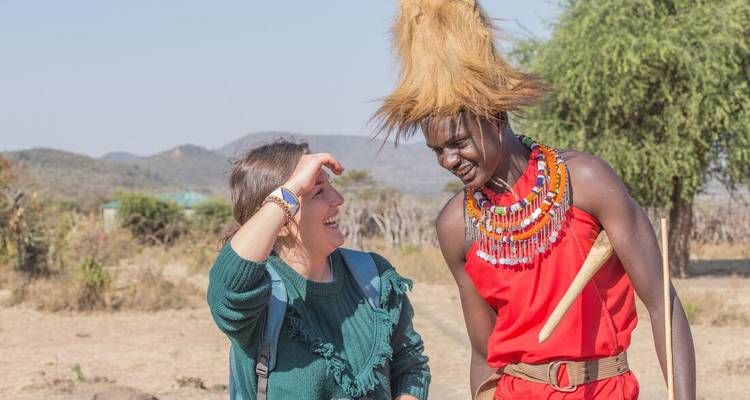 A traveller laughs with a traditionally dressed Maasai man under bright sun on open countryside.