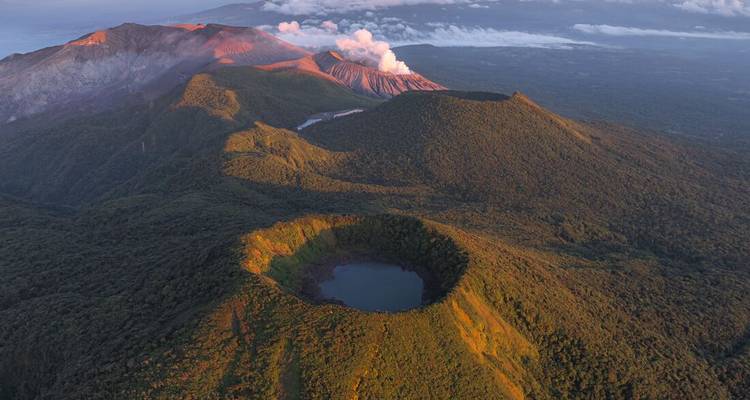 Aerial sunrise view of volcanic craters and lush green ridges with distant steam plume.