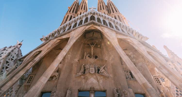 Vue en contre-plongée des statues sculptées de la façade de la Passion de la Sagrada Familia sur un ciel dégagé avec halo solaire.