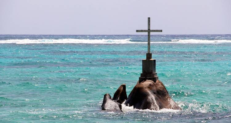 A lone cross stands atop a granite outcrop in bright turquoise ocean swells