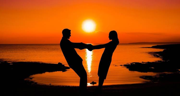 Silhouetted couple holding hands against a fiery orange sunset on the coast