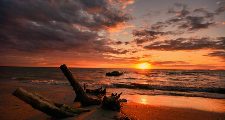 Ein dramatischer Sonnenuntergang färbt den Himmel über einem ruhigen tropischen Strand mit Treibholz und einem kleinen Boot auf sanften Wellen orange.