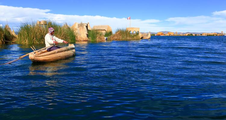 Un hombre local rema una canoa de junco a través de las aguas azul profundo del Lago Titicaca con islas flotantes atrás.