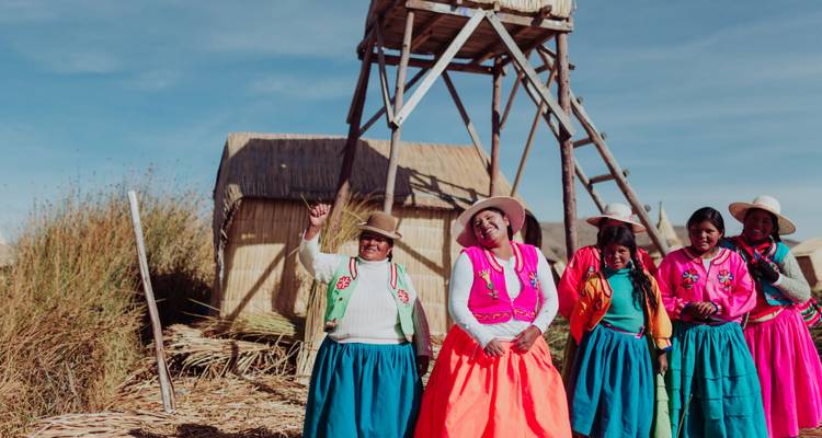 Mujeres con vestidos tradicionales de colores brillantes cantan y hacen gestos junto a cabañas de junco y una torre de vigilancia de madera en el Lago Titicaca.