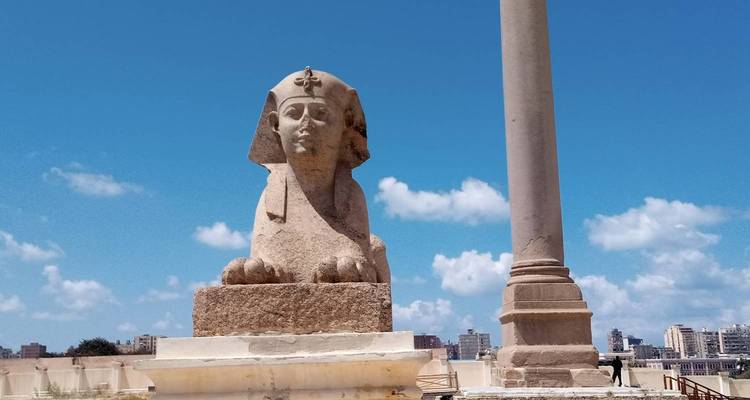 Ancient sphinx statue beside Pompey's Pillar against a bright blue sky and city skyline.