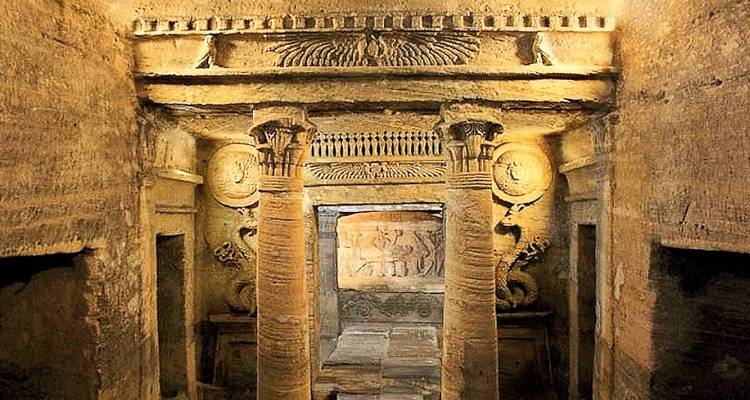 Underground catacomb chamber with carved columns, reliefs and dim golden lighting.