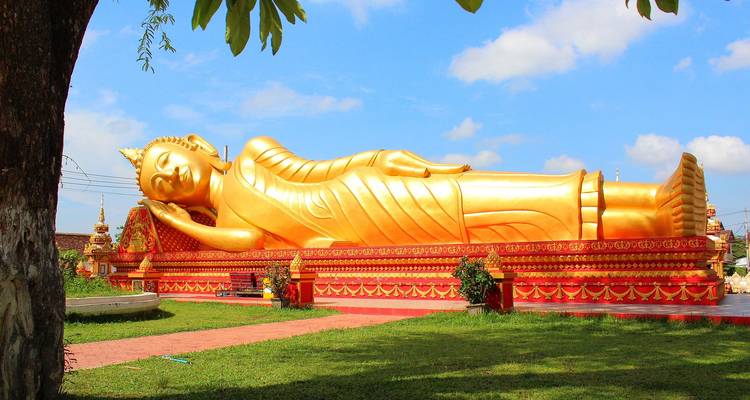 Große goldene liegende Buddha-Statue ruht im Freien auf einem roten Sockel inmitten von grünem Gras und blauem Himmel.