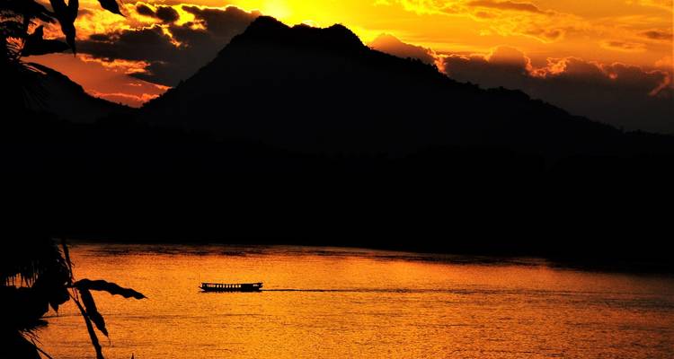 Silhouetten zerklüfteter Berge ragen über einem ruhigen Fluss empor, wo ein einsames Longtail-Boot unter einem feurig orangefarbenen Sonnenuntergang dahintreibt.