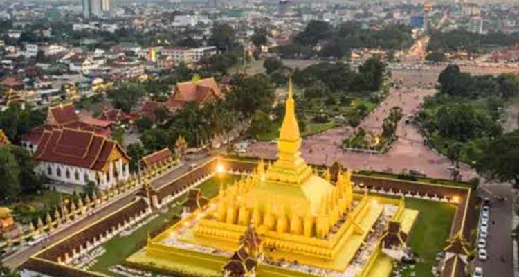 Luftaufnahme in der Abenddämmerung der beleuchteten goldenen Pha-That-Luang-Stupa, umgeben von der Stadtlandschaft.