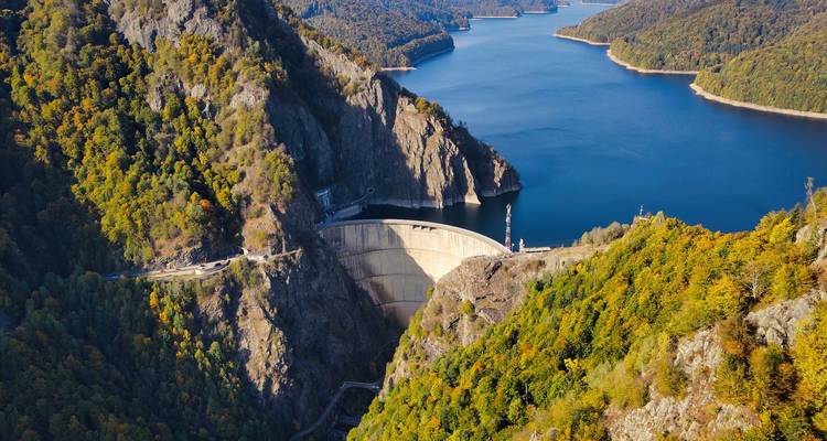 Vue aérienne panoramique d'un grand barrage niché entre des montagnes boisées et un réservoir d'un bleu profond.