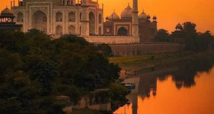 El Taj Mahal brillando en la luz naranja del atardecer con un río tranquilo reflejando el monumento y los árboles cercanos.