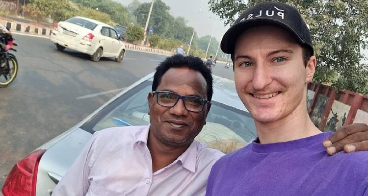 Two men smile for a roadside selfie beside a parked silver car on a city street.