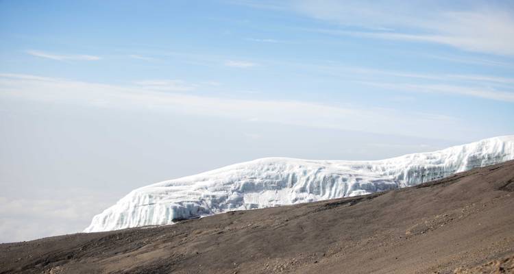Paroi de glacier blanc perchée sur le versant de couleur cendre du Kilimandjaro sous un ciel bleu.