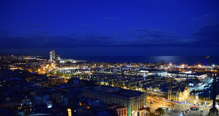 Nighttime aerial of Barcelona’s waterfront and marina with city lights reflecting on the Mediterranean.