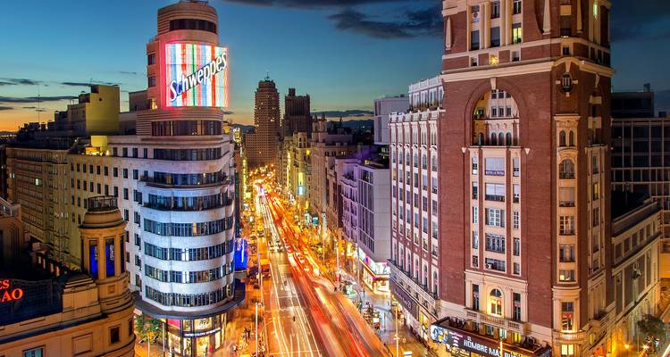 Dusk view over Madrid’s Gran Vía with the neon Schweppes sign and light trails of traffic.