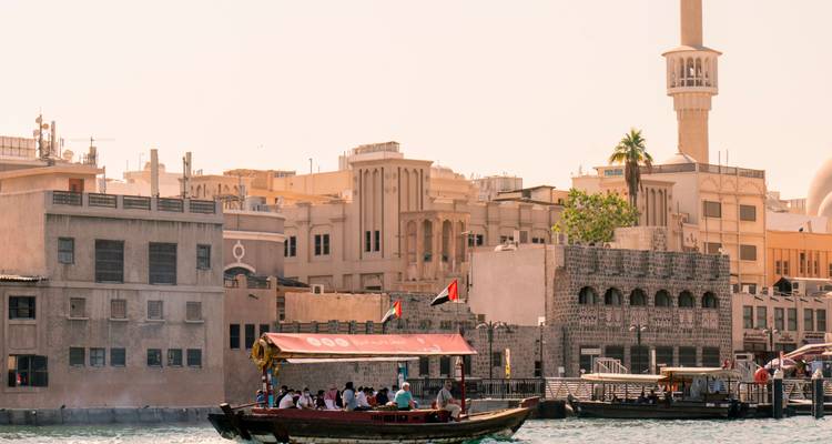 Wooden passenger boat full of commuters crossing a sun-washed creek lined with sand-colored Middle Eastern architecture and a minaret.