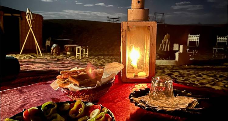 Une lanterne brille sur une table basse avec du pain, des poivrons et des sables désertiques en arrière-plan la nuit.