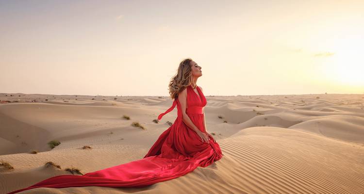 Une femme vêtue d'une robe rouge fluide s'agenouille avec grâce sur les vagues douces du sable au coucher du soleil.