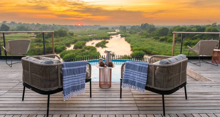 Vue du lever de soleil depuis une terrasse en bois avec des fauteuils et des serviettes donnant sur une rivière sinueuse et un ciel vibrant.