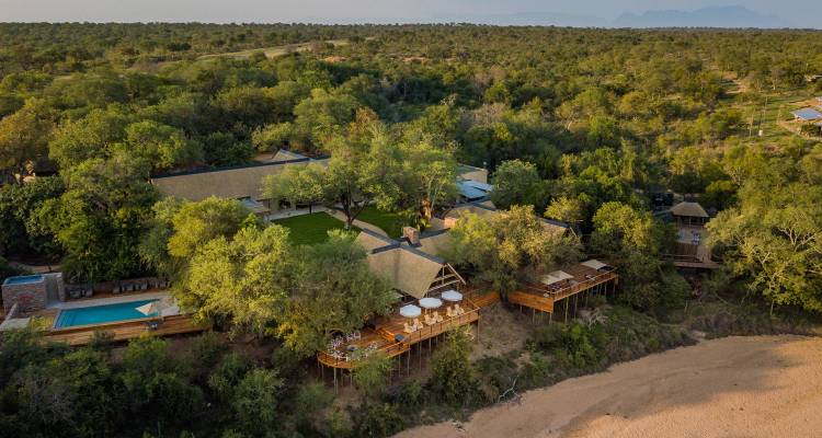 Vue aérienne d'un complexe de lodge de safari spacieux abrité par des arbres denses sur les rives d'une rivière en savane buissonnante.