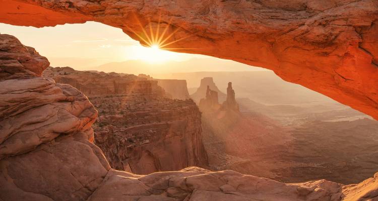 Sunburst shining through Mesa Arch framing crimson desert canyons below.
