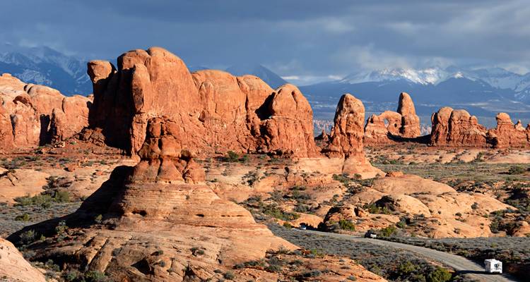 Dramatic red sandstone formations rise under stormy skies in Arches National Park with snowy mountains beyond.