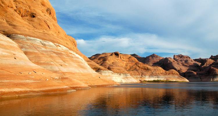 Striped orange and white canyon walls meet the calm reflective waters of Lake Powell under blue sky.