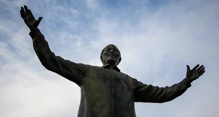 Statue de bronze de Nelson Mandela les bras levés devant un ciel bleu et blanc.